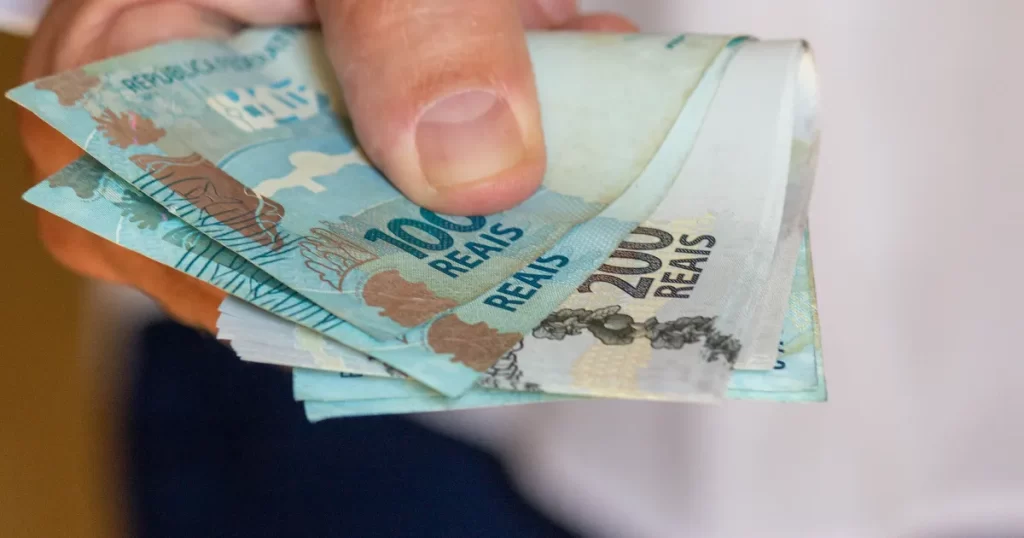Mão segurando notas de reais de 100 e 200, simbolizando a luta para sair das dívidas e gerenciar finanças pessoais.