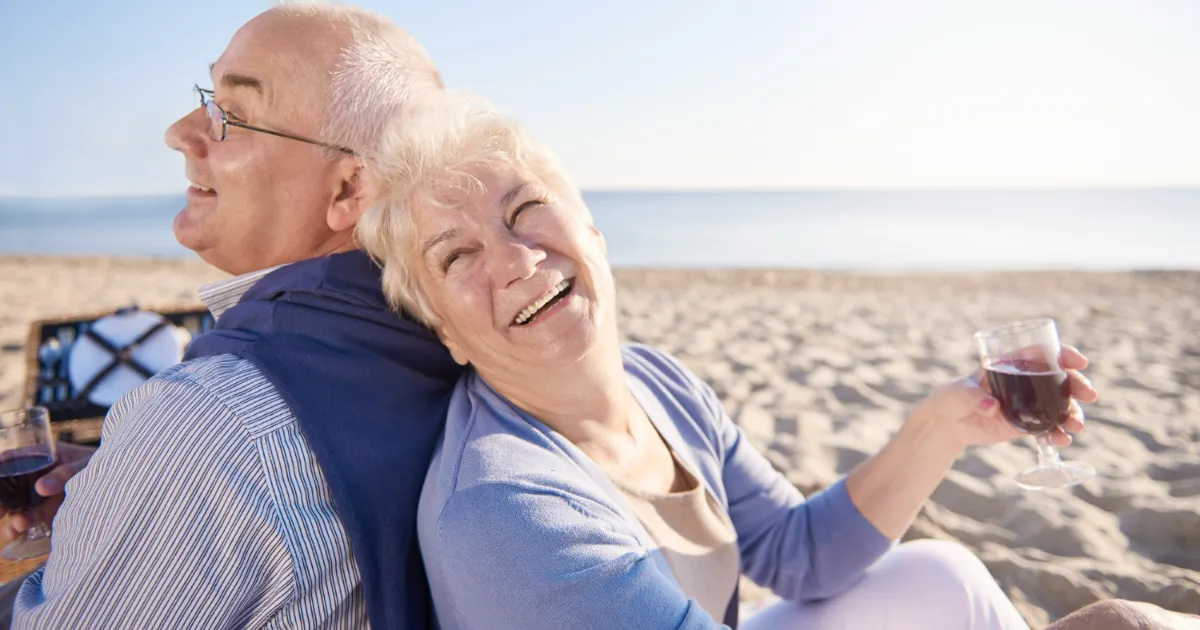 Um casal de idosos feliz e relaxado na praia, o homem de costas e a mulher rindo enquanto segura uma taça de vinho, simbolizando a realização financeira e a tranquilidade na aposentadoria.