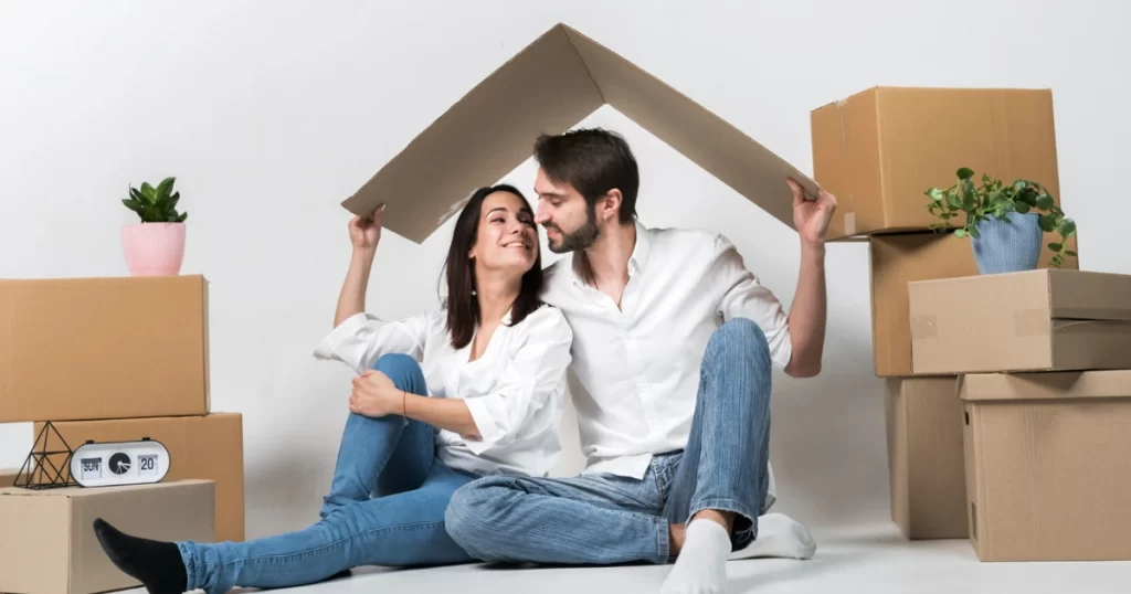 Casal sorridente sob um telhado de papelão, cercado por caixas de mudança, representando a construção de um futuro financeiro seguro com investimento em imóveis, como os FIIs.