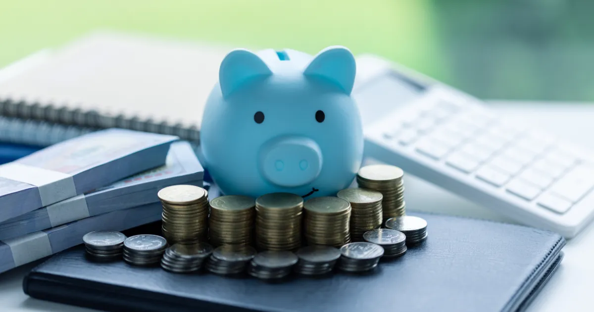 Cofrinho azul em forma de porquinho com moedas e pilhas de dinheiro ao redor, simbolizando a economia e o investimento seguro em renda fixa.