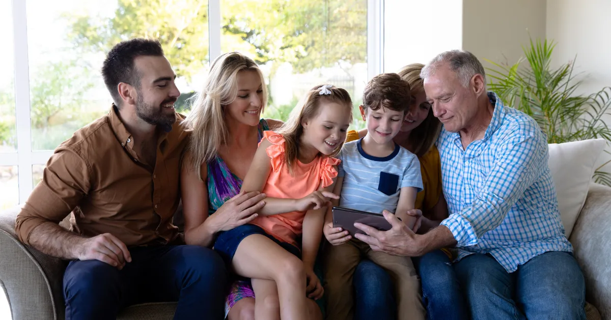 Uma família multigeneracional sorrindo enquanto olha para um tablet, representando as diferentes fases da vida e a necessidade de diversificação de investimentos por idade.