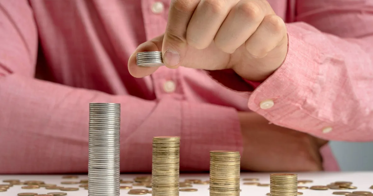 Pessoa empilhando moedas para representar o crescimento dos investimentos conforme diferentes indexadores de renda fixa, como CDI, Selic e IPCA.