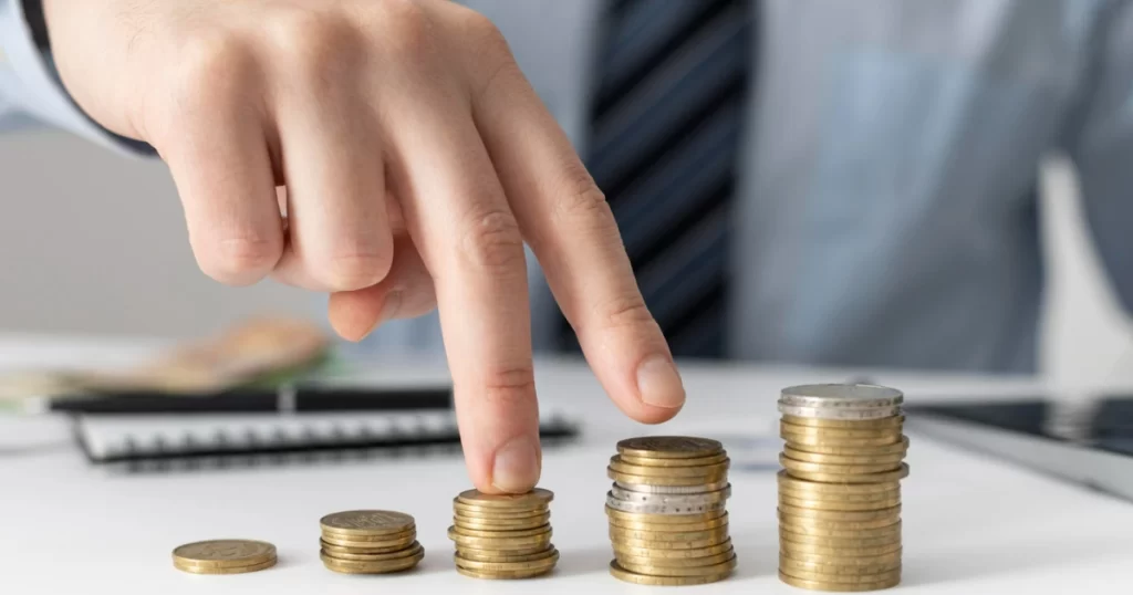 Mão de uma pessoa empilhando moedas, com uma seta vermelha de crescimento ao fundo, simbolizando a acumulação de patrimônio e a valorização de investimentos em renda fixa.