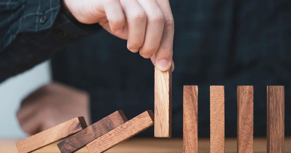 Uma mão posiciona um bloco de madeira para parar a queda de uma sequência de dominós, simbolizando o stop loss como uma barreira de proteção contra perdas.