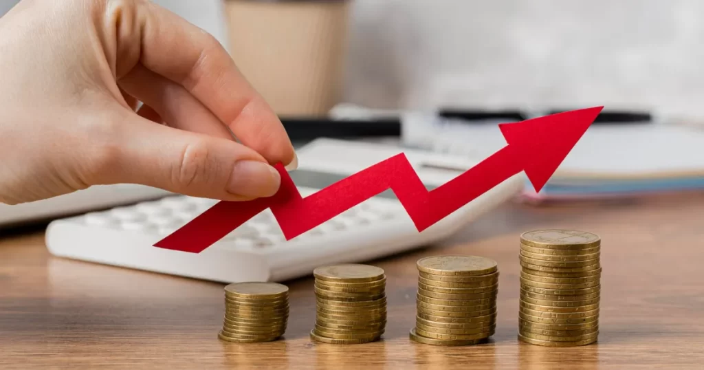 Mão posicionando uma seta de crescimento vermelha sobre moedas empilhadas, simbolizando o aumento de dividendos e o sucesso do investimento na B3.