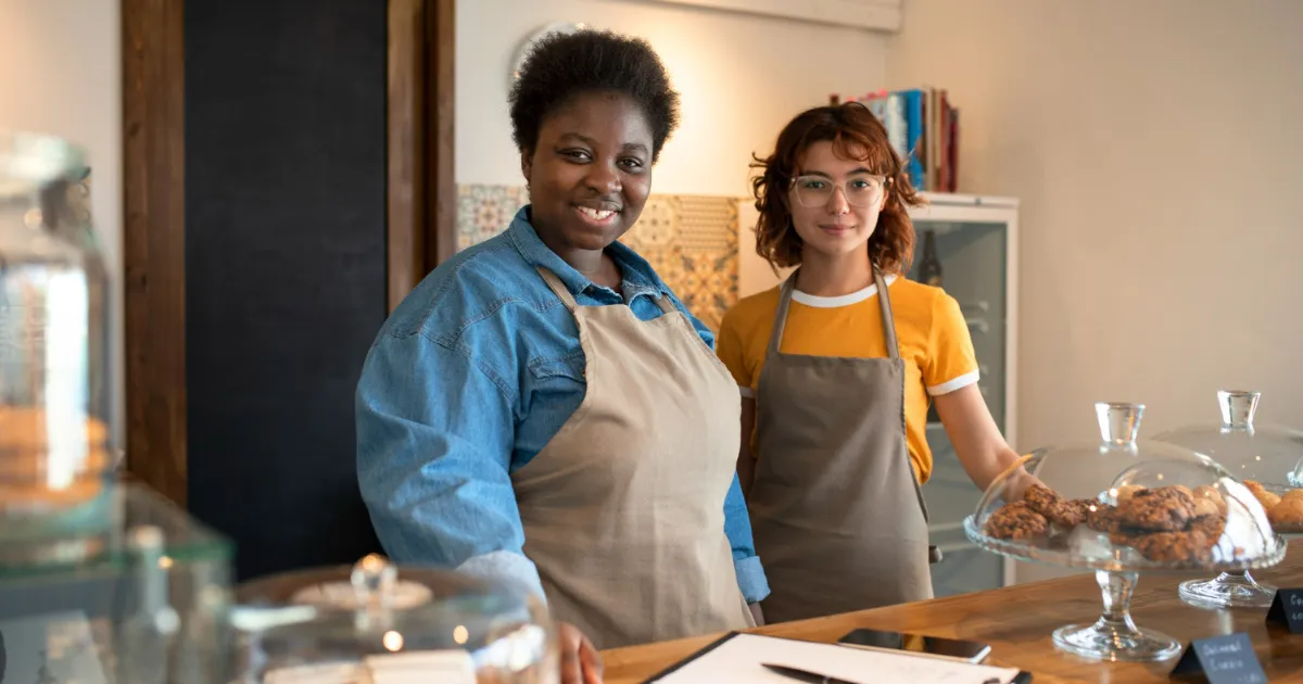 Duas mulheres sorrindo atrás do balcão de uma cafeteria ou padaria, usando aventais, representando proprietárias ou funcionárias de uma franquia de alimentação.