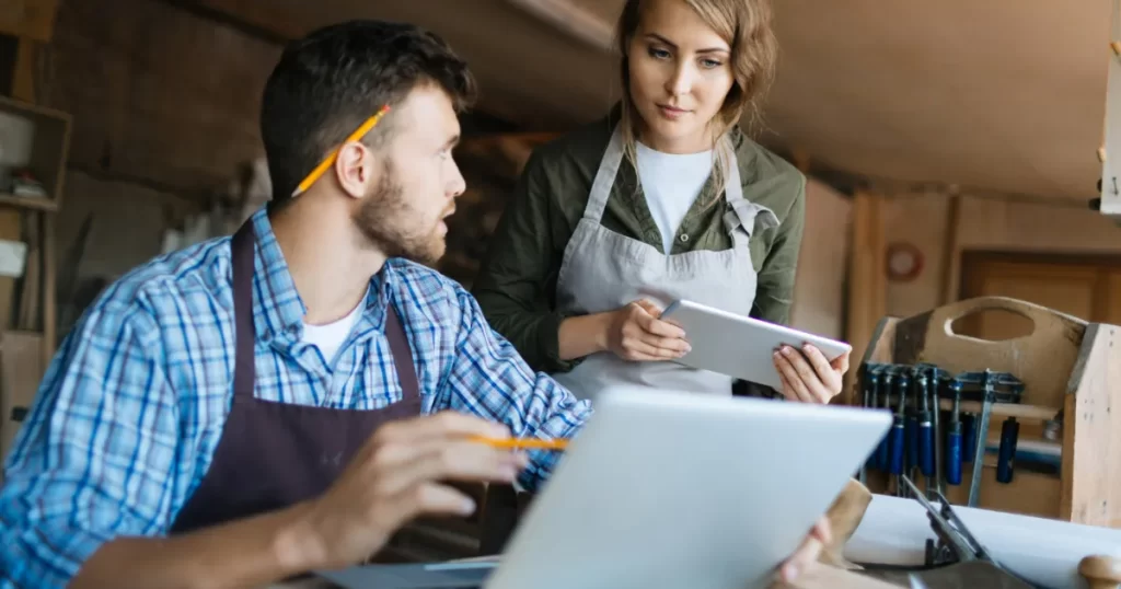Dois empreendedores, um homem e uma mulher, em uma oficina ou escritório, usando um laptop e um tablet para gerenciar seu negócio, simbolizando o trabalho em equipe e a eficiência.