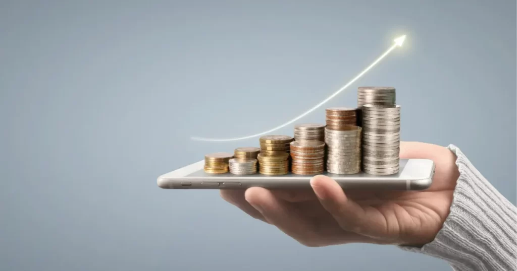 Mão segurando um smartphone com moedas empilhadas em forma de gráfico de crescimento, simbolizando o monitoramento de dividendos e JCP através de plataformas digitais.