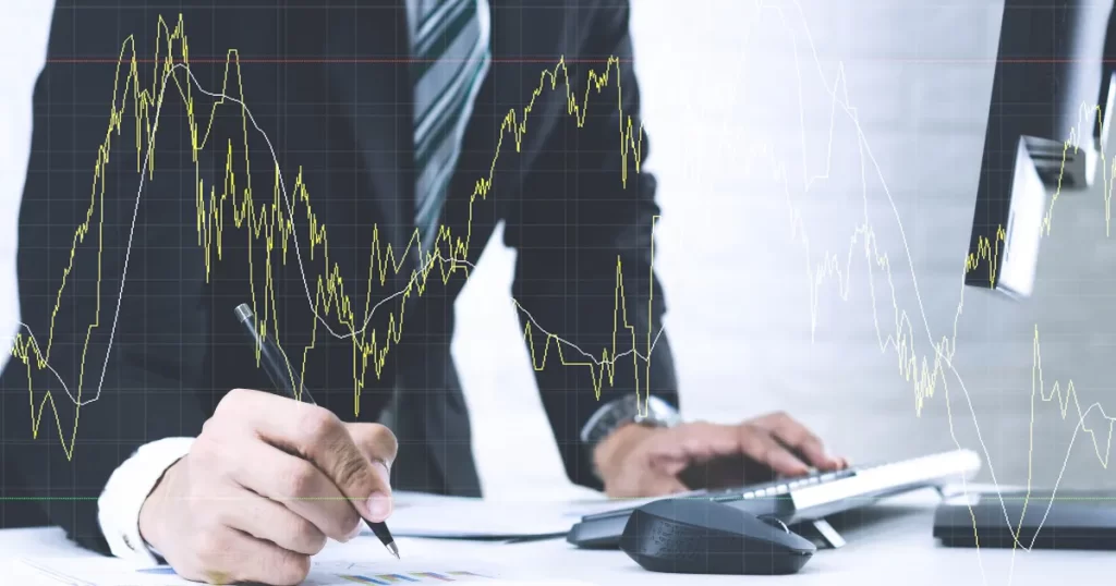 Um homem de terno calculando em uma mesa com gráficos e um monitor ao fundo exibindo um gráfico de ações, representando o processo de calcular o valor justo de uma ação no valuation.