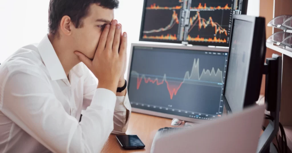 Jovem trader estressado e pensativo, com as mãos no rosto, analisando gráficos financeiros em dois monitores, que exibem padrões de preço em alta e baixa.