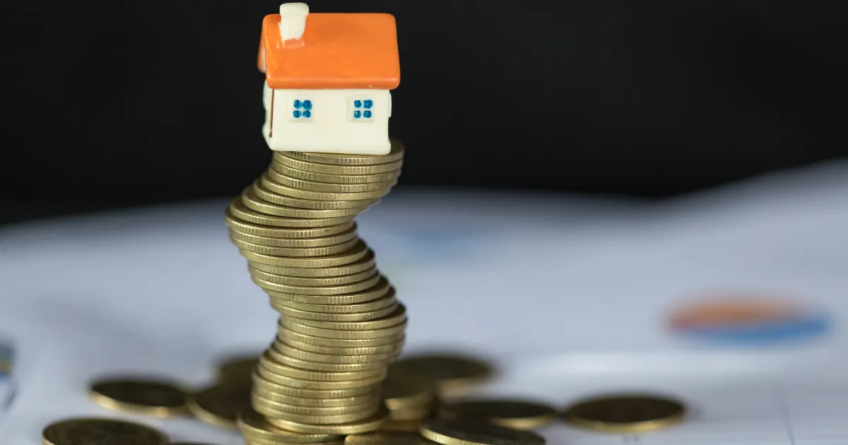 Uma casa em miniatura equilibrada sobre uma pilha de moedas de ouro em cima de documentos financeiros, simbolizando a união entre o mercado imobiliário e os investimentos.
