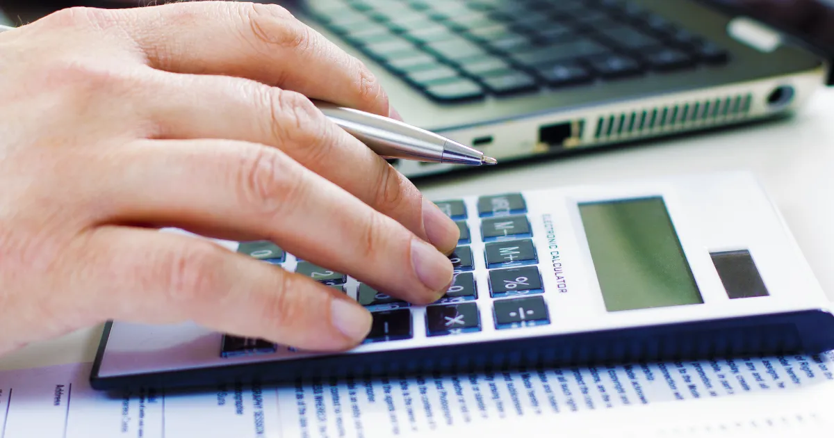 Mão usando uma calculadora e uma caneta, com um laptop ao fundo, simbolizando a gestão e o controle financeiro essenciais para um negócio.
