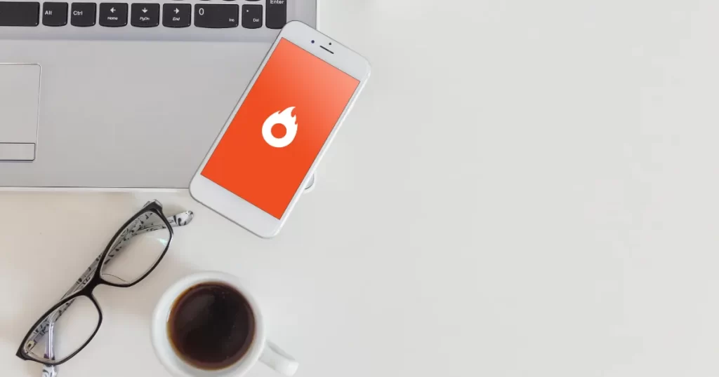 Uma imagem de topo de uma mesa de trabalho com um laptop, óculos e um smartphone exibindo o logo da Hotmart, simbolizando o trabalho de afiliado digital.