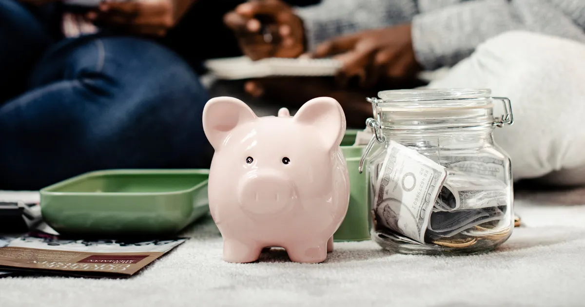 Um cofrinho de porco rosa ao lado de um pote de vidro com notas e moedas, simbolizando a economia para investir com pouco dinheiro.