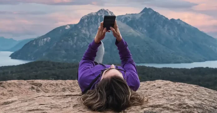 Mulher deitada em uma rocha, usando o celular com um cenário de montanhas e lago ao fundo, simbolizando a liberdade e a tranquilidade de viajar o mundo após quitar as dívidas.