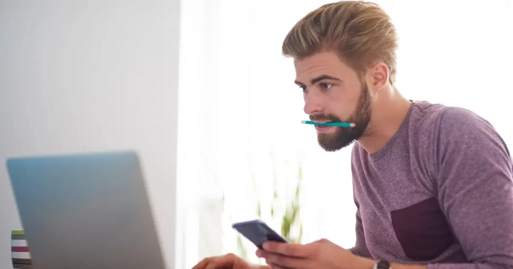 Homem com barba e um lápis na boca, concentrado em um laptop e segurando um smartphone, simbolizando um profissional freelancer trabalhando em home office.