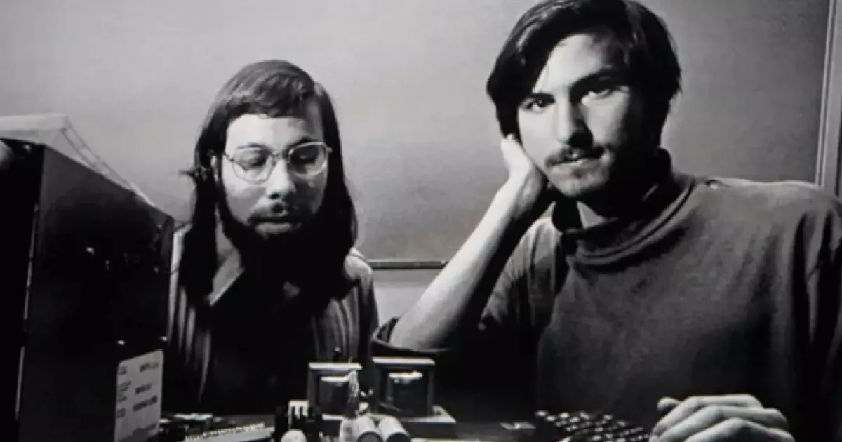 Foto em preto e branco de Steve Jobs e Steve Wozniak, os co-fundadores da Apple, sentados em uma mesa com equipamentos de computador nos primórdios da empresa.